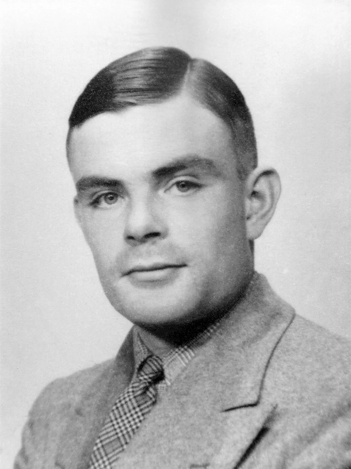 A black and white bust photograph of Alan Turing. He is looking dead on at the camera, and his hair is slicked to the side. He is wearing a suit, a button up, and a tie. The background is white.