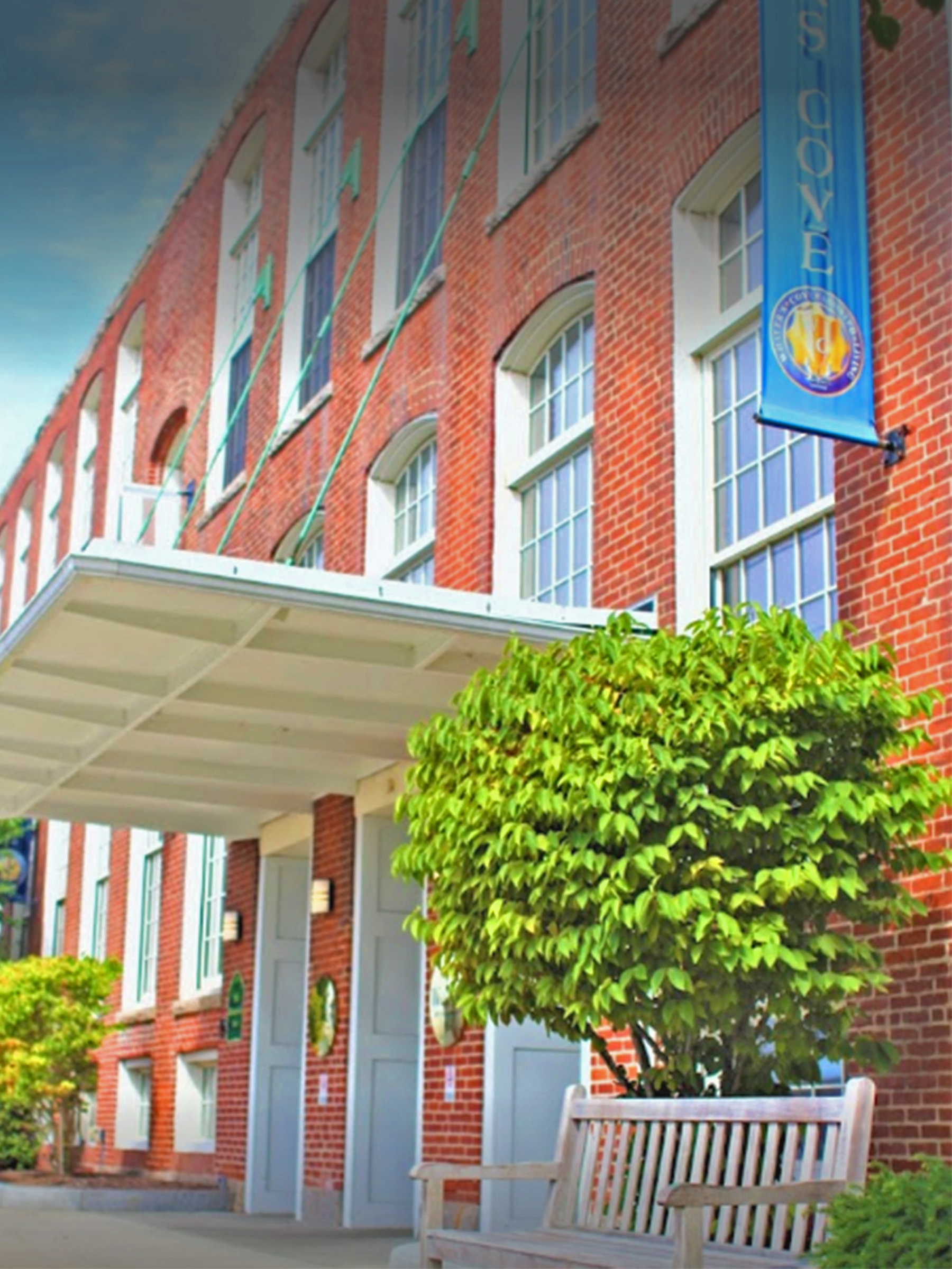 Entrance to a red brick building with big white windows, a bench, bushes and an overhang above the door.