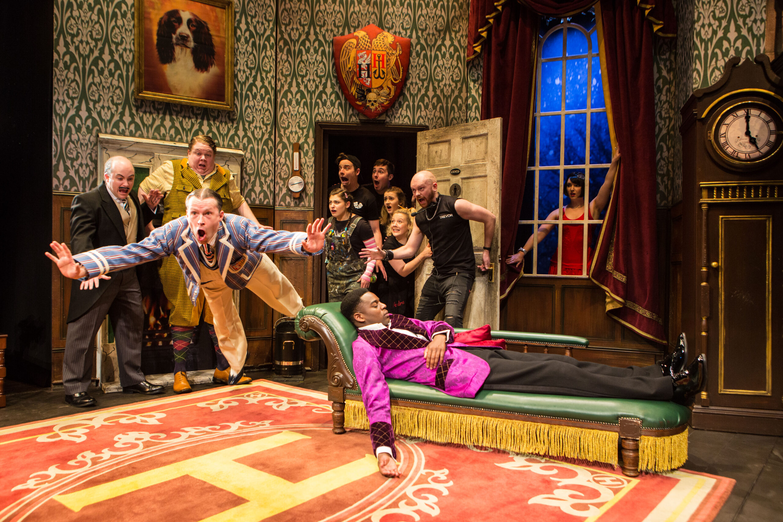 a man in blue falling as the stage crew opens the door. a man in purple lays unconscious on the couch, a girl in a red dress stands outside the window, and 2 men (one in yellow, one in black with a mustache) stand shocked beside him