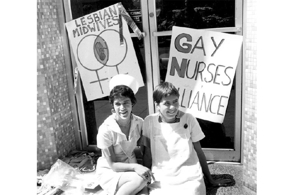 Old black and white image of two nurses sitting in front of two signs one saying "Gay Nurses Alliance" and the other saying "Lesbian Midwives"