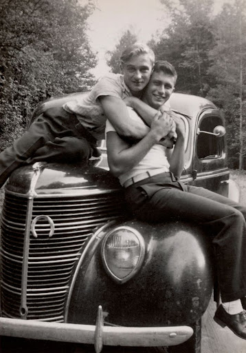 A photograph found by Nini and Treadwell. Shows two men sitting on a car, one wrapping his arms around the other while the other holds onto his arms. They are smiling at the camera.