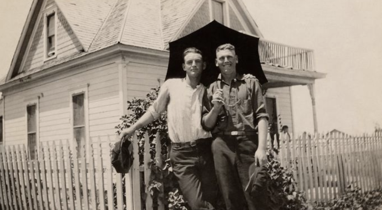 A photograph of two men under an umbrella. One is smiling while the other has a settled look on his face as he holds the shoulder of the other, who is holding up the umbrella. They are infront of a house and leaning on a fence.