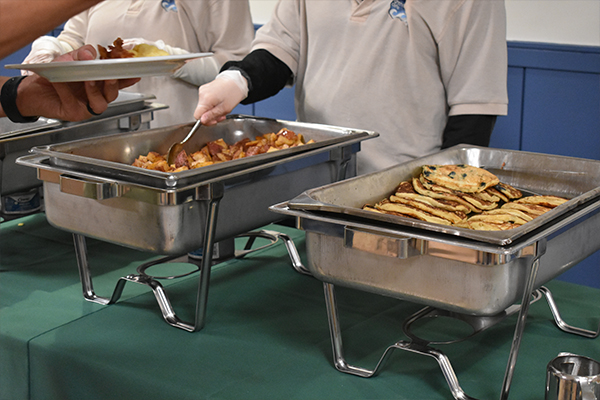 Two people serving potatoes and pancakes out of a metal heating tray.