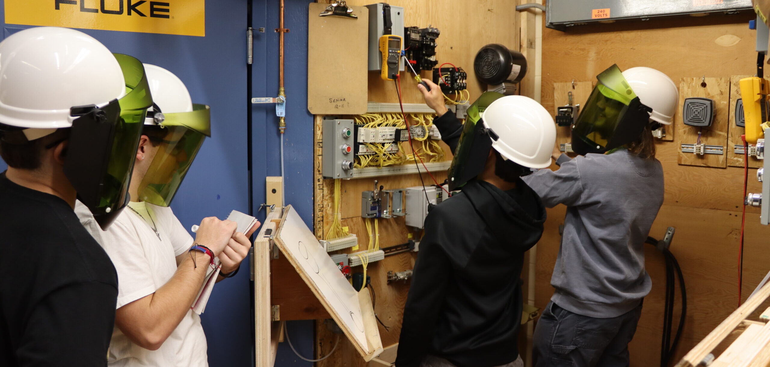 Two students working on an electrical project while two other students watch from the sidelines and take notes. 