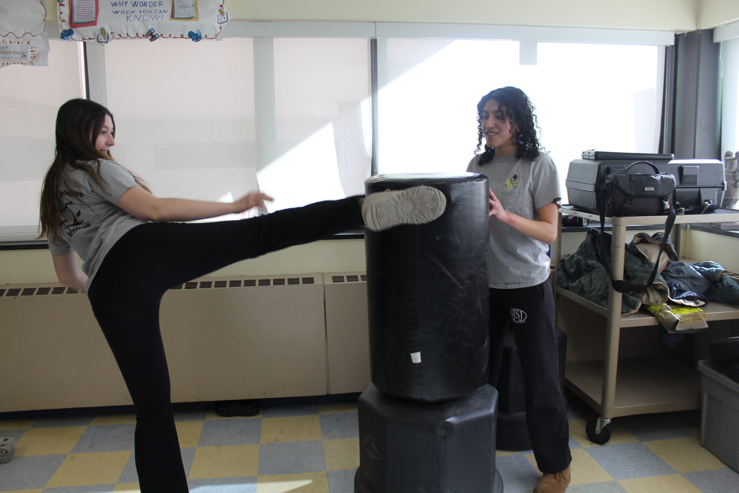 Two students practicing physical combat. One student is kicking a punching bag, and the other student holds it still. 